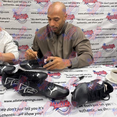 Man signing black Puma soccer cleats at a signing event with an AllStar Signings backdrop.