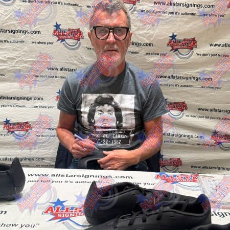 Autograph session: man wearing glasses and a gray tee sits at a signing table in front of an AllStar Signings backdrop, holding a marker with black sneakers displayed on the table.