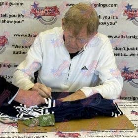 Older man in a white Adidas jacket signs a navy sports jersey at an All-Star Signings table, with a branded backdrop in the background.