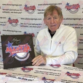 Older man in a white zip-up jacket sits at a table with a large AllStar Signings signboard featuring the logo and a signature, against a backdrop of AllStar Signings banners.