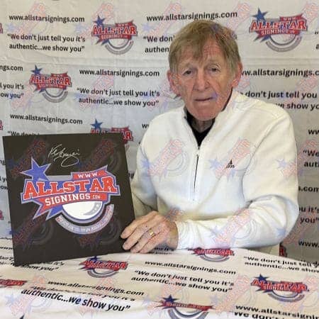 Older man in a white zip-up jacket sits at a table with a large AllStar Signings signboard featuring the logo and a signature, against a backdrop of AllStar Signings banners.