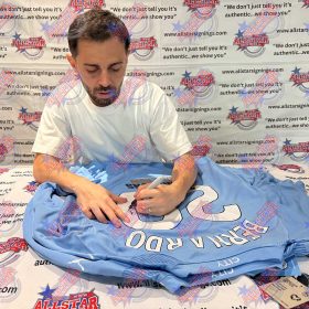 Man in a white T-shirt signs a light blue sports jersey at a signing event; backdrop shows All-Star Signings logos.