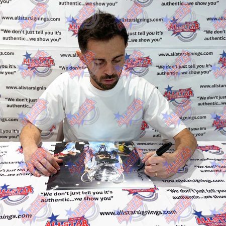Man in a white T-shirt signs a glossy photo at a signing event, seated at a table with an All Star Signings backdrop in the background.