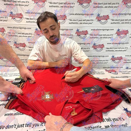 Man signing a red Portugal soccer jersey at a signing event, with hands holding the jersey and a branded backdrop behind.