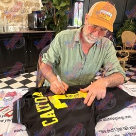 Man in an orange cap signs a black T‑shirt with a yellow graphic at a signing event.