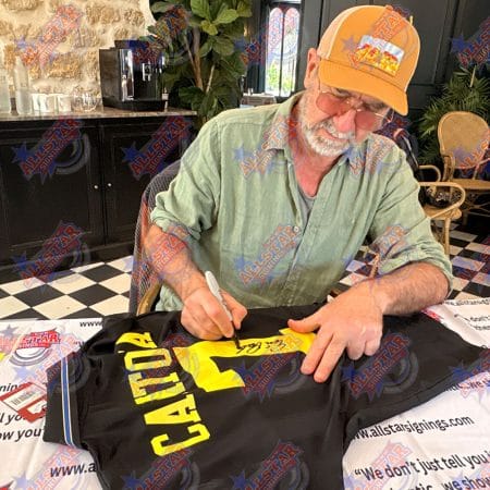 Man in an orange cap signs a black T‑shirt with a yellow graphic at a signing event.